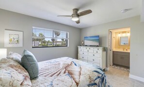Ein Schlafzimmer mit einem Bett, einer Kommode, einem Deckenventilator, einem Fenster mit Jalousien, einem Fernseher und einem Badezimmer mit Waschtisch und Spiegel.