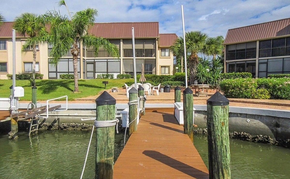 A wooden dock leading to a building with a red roof, surrounded by palm trees and greenery.