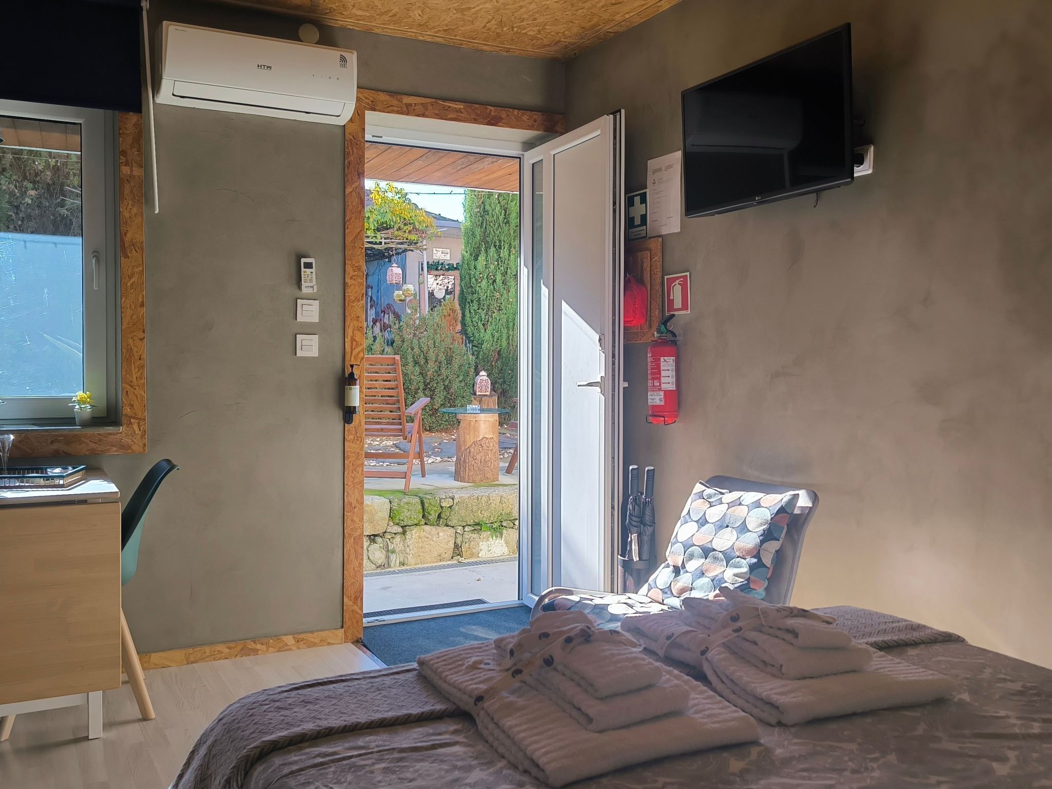 Une chambre d’hôtel équipée d’un lit, d’un bureau, d’une chaise, d’une télévision et d’une porte-fenêtre coulissante donnant sur un espace extérieur.