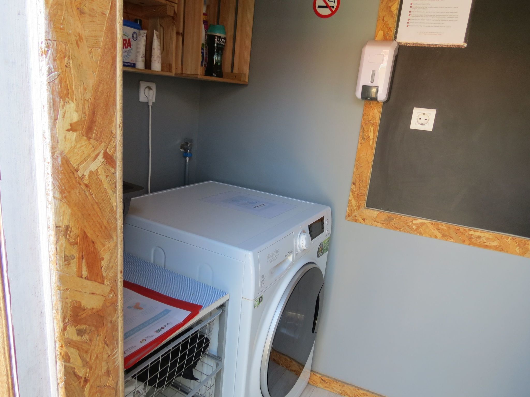 Une petite buanderie comprenant un lave-linge, un étendoir et une petite étagère sur laquelle sont rangés divers objets.
