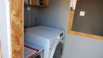 Une petite buanderie comprenant un lave-linge, un étendoir et une petite étagère sur laquelle sont rangés divers objets.