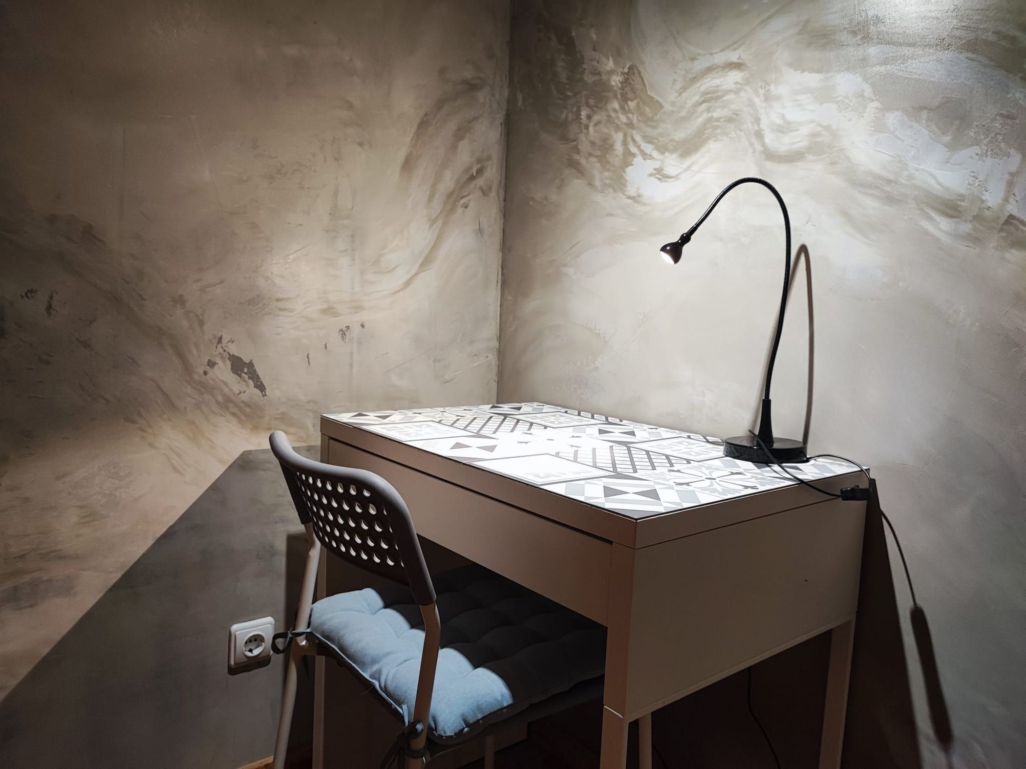 Un petit bureau carré, surmonté d’une nappe à motifs géométriques, une chaise noire avec un dossier en tissu grillagé et une seule lampe de bureau noire.