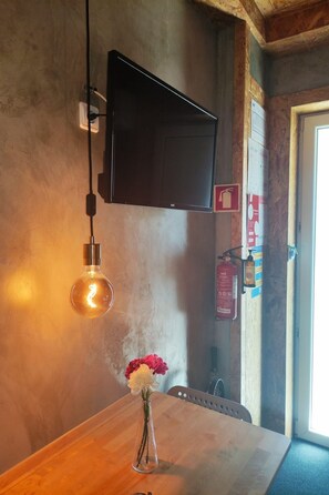 Een kamer met een houten tafel, een bloemenvaas, een hangende lamp, een bevestigde televisie en een brandblusser aan de muur.