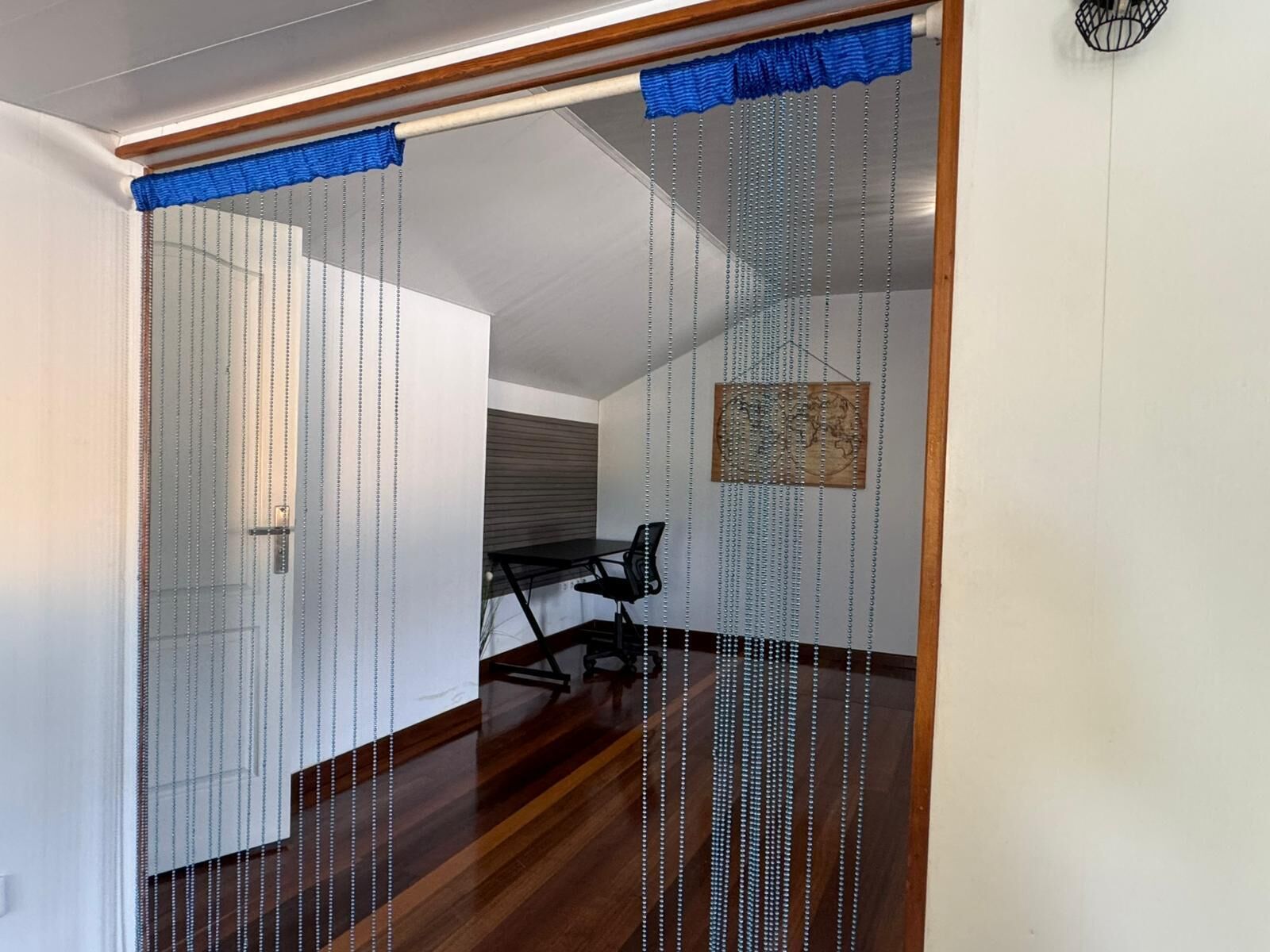 Quarto com piso de madeira, uma escrivaninha e uma cadeira. Há uma porta deslizante com cortina azul.
