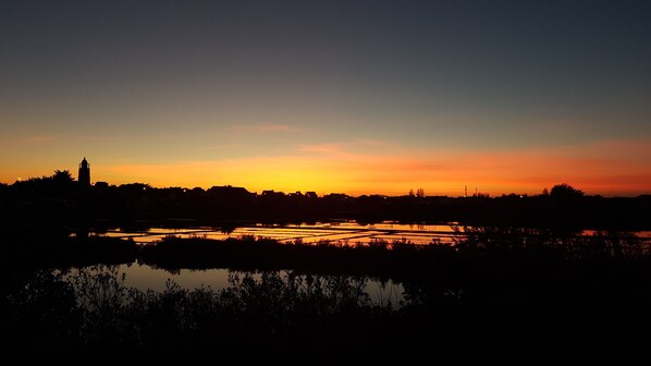 A sunset over a landscape with water reflecting the sky, silhouettes of buildings, and a tower structure.