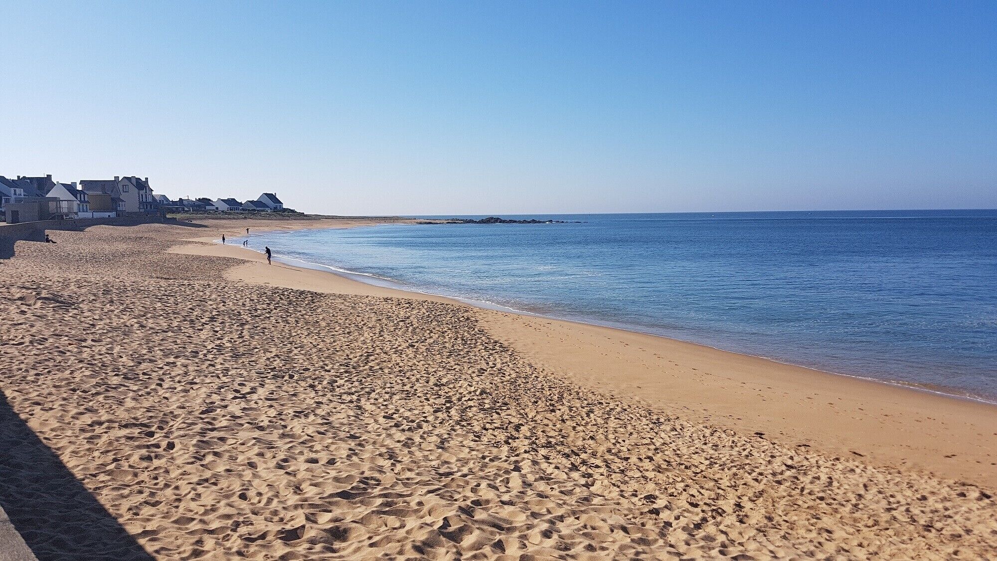 Una playa arenosa con huellas, un cielo azul despejado y edificios al fondo.