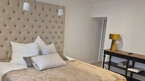 Um quarto com cabeceira estofada, cama grande com várias almofadas, mesa de cabeceira com abajur e um banco com almofadas.