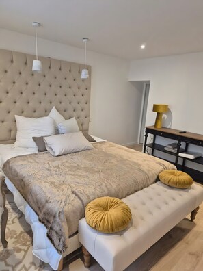 A bedroom with a tufted headboard, a large bed with multiple pillows, a bedside table with a lamp, and a bench with cushions. - Exceptional Apartment 500m from Beach with Wi-Fi (Batz-sur-Mer)