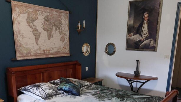 Uma cama de madeira com colcha florida, um mapa-múndi, um retrato e uma mesa pequena com abajur.