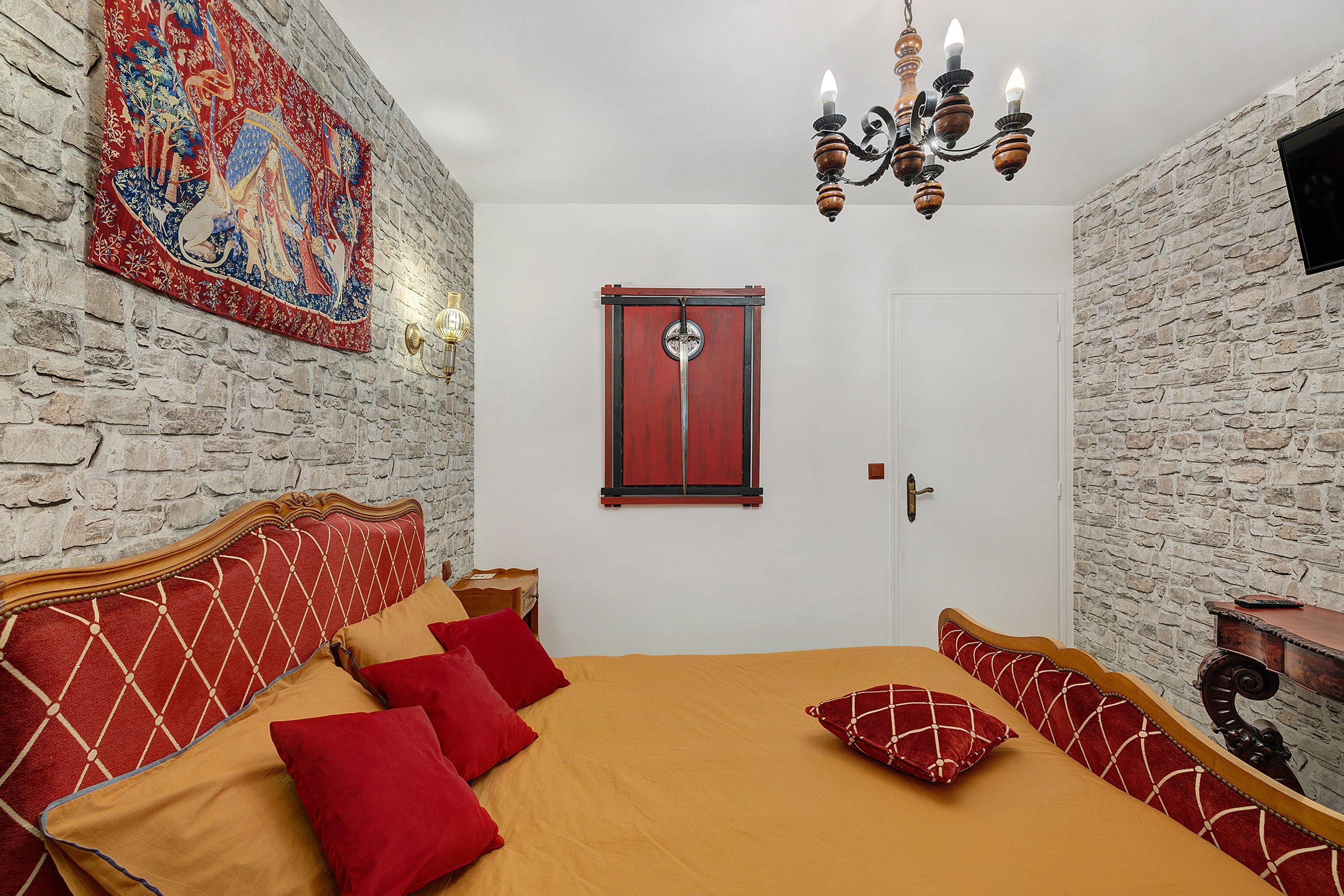 Une pièce avec un mur en pierre, une tapisserie, un lit en bois avec une literie rouge et beige, et un lustre.