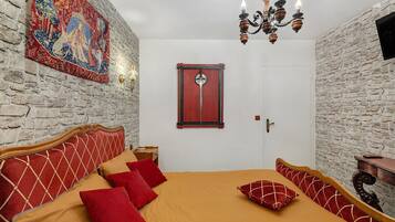 Une pièce avec un mur en pierre, une tapisserie, un lit en bois avec une literie rouge et beige, et un lustre.