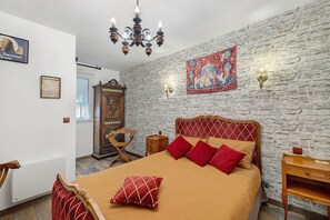 A bedroom with a stone wall, a bed with red and beige bedding, a wooden nightstand, a wooden chair, a chandelier, and a tapestry.