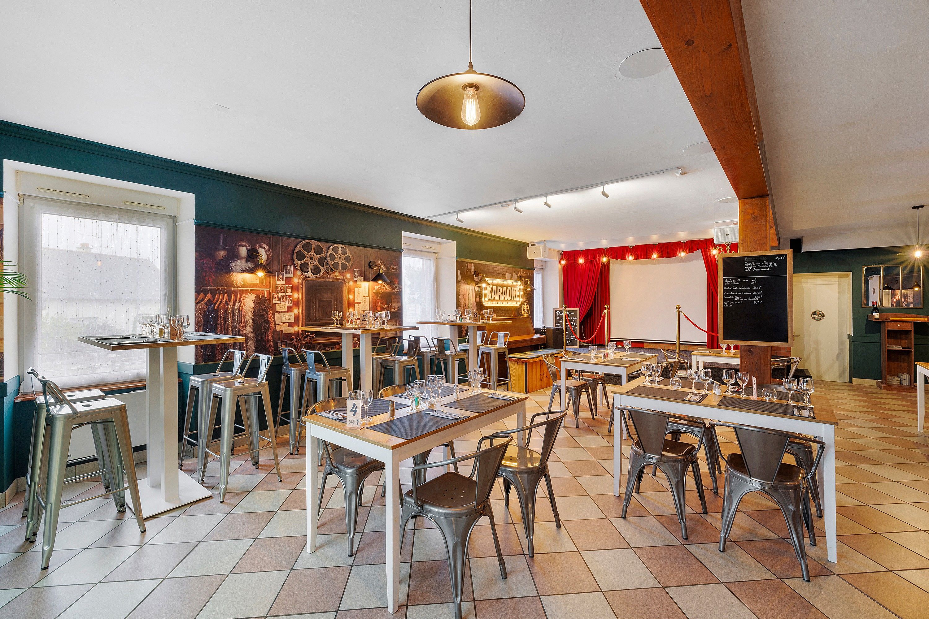 Un restaurant avec de nombreuses tables et chaises, une scène surmontée d’un rideau rouge et un menu écrit à la craie sur un tableau noir.