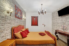 Une chambre avec un mur en pierre, un lit avec une literie rouge et beige, un lustre et une table de chevet en bois.