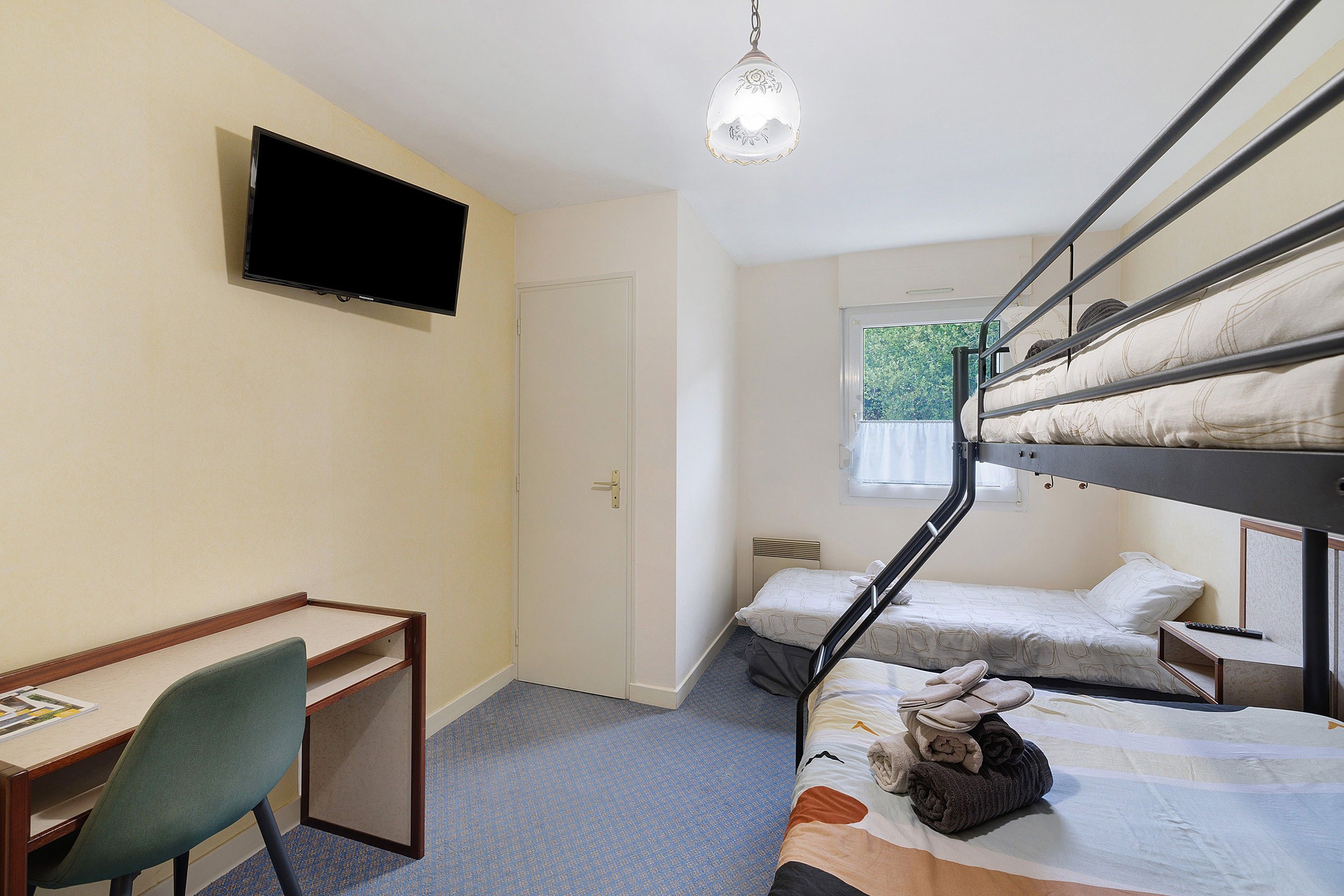 Une chambre de dortoir équipée de lits superposés, d’un bureau, d’une chaise, d’un téléviseur et d’une fenêtre donnant sur un paysage verdoyant.