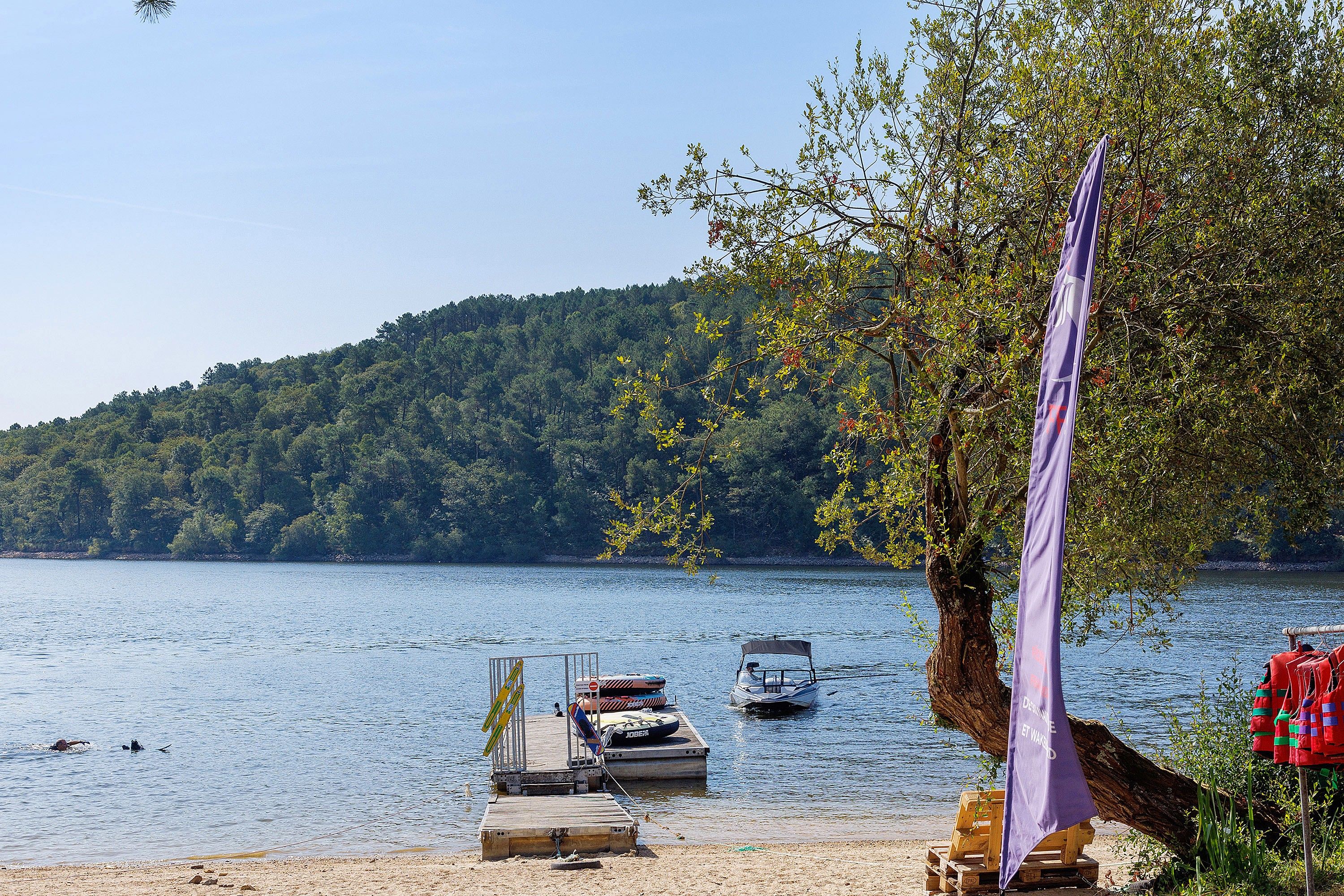 Une jetée au bord du lac avec des bateaux, une plateforme en bois et, au loin, une colline boisée.