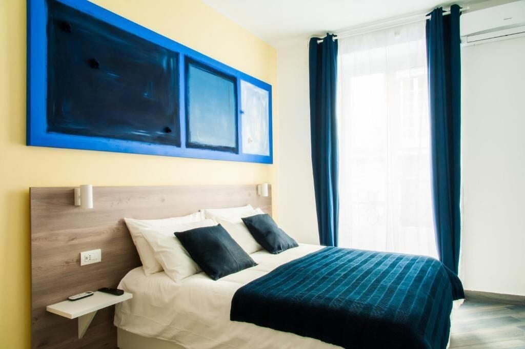 A hotel room with a bed, a nightstand, a wall-mounted artwork, and a window with curtains.