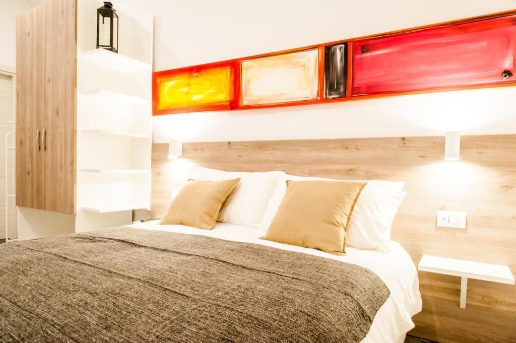 Quarto de hotel com cabeceira de madeira, cama com roupa de cama branca e almofadas bege, manta cinza e prateleira fixa com luzes vermelhas e amarelas.