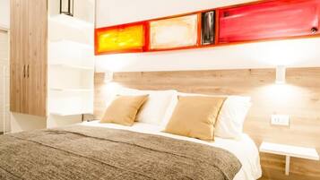 Quarto de hotel com cabeceira de madeira, cama com roupa de cama branca e almofadas bege, manta cinza e prateleira fixa com luzes vermelhas e amarelas.