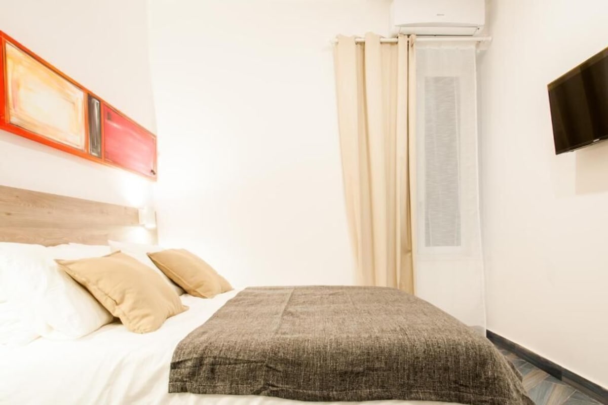 A hotel room with a bed, pillows, a flat-screen TV, an air conditioning unit, and a window with curtains.