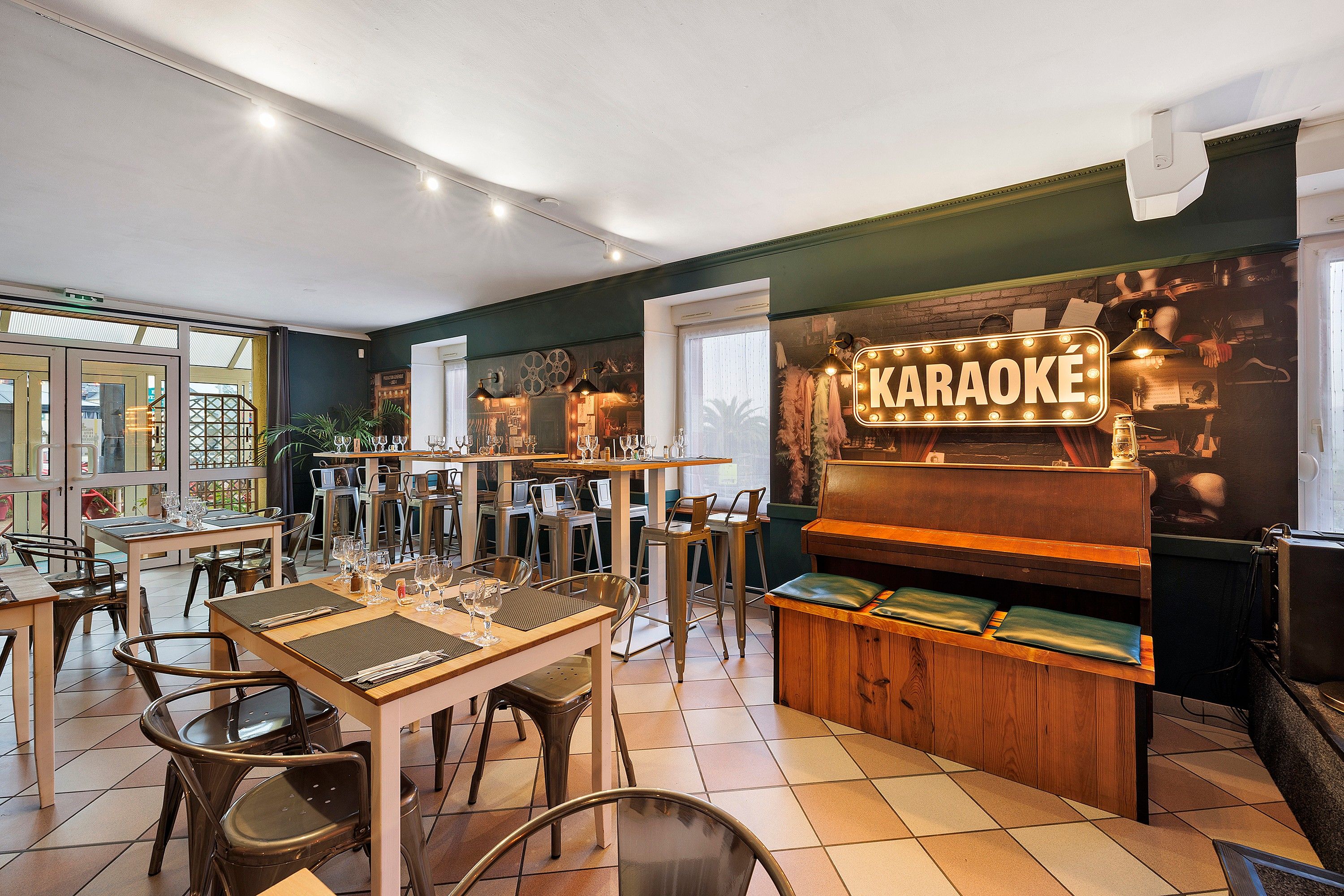 Un bar karaoké avec des tables et des chaises, une enseigne indiquant « KARAOKÉ », et une scène équipée d’instruments de musique.