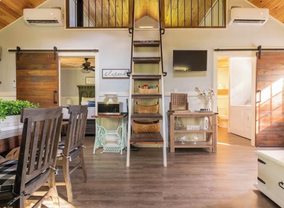 Une pièce spacieuse dotée d’un plancher en bois, d’un escalier central, d’un coin repas avec des chaises en bois et d’un téléviseur fixé au mur.