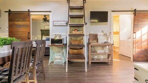 Een ruime kamer met een houten vloer, een centrale trap, een eethoek met houten stoelen, en een aan de muur bevestigde televisie.