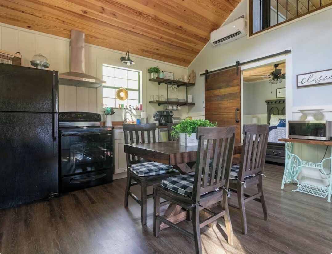 Une cuisine avec un réfrigérateur noir, une cuisinière et un plafond en bois. On y trouve une table à manger avec des chaises et une plante.