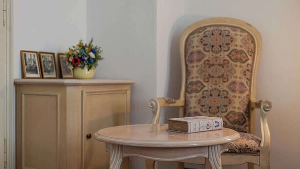 A room with a round table, a chair with a patterned cushion, a cabinet with framed pictures, and a vase with flowers. - The Royal Hlavna - Balcony Apartment (Old Town, Košice)