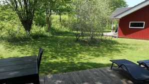 A deck with two lounge chairs and a wooden table, surrounded by a grassy yard and a red building. - Helles Kustenhaus mit Sauna (Grossenbrode)