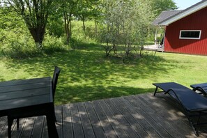 A deck with two lounge chairs and a wooden table, surrounded by a grassy yard and a red building. - Helles Kustenhaus mit Sauna (Grossenbrode)