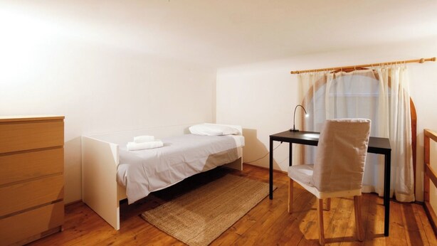 Ein kleines Zimmer mit einem Einzelbett, einem Schreibtisch, einem Stuhl, einer Holzkommode und einem Fenster mit Vorhängen.