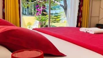 Habitación de hotel con una cama, una lámpara roja sobre una mesita de noche de madera y vista a un jardín a través de una puerta corrediza de vidrio.