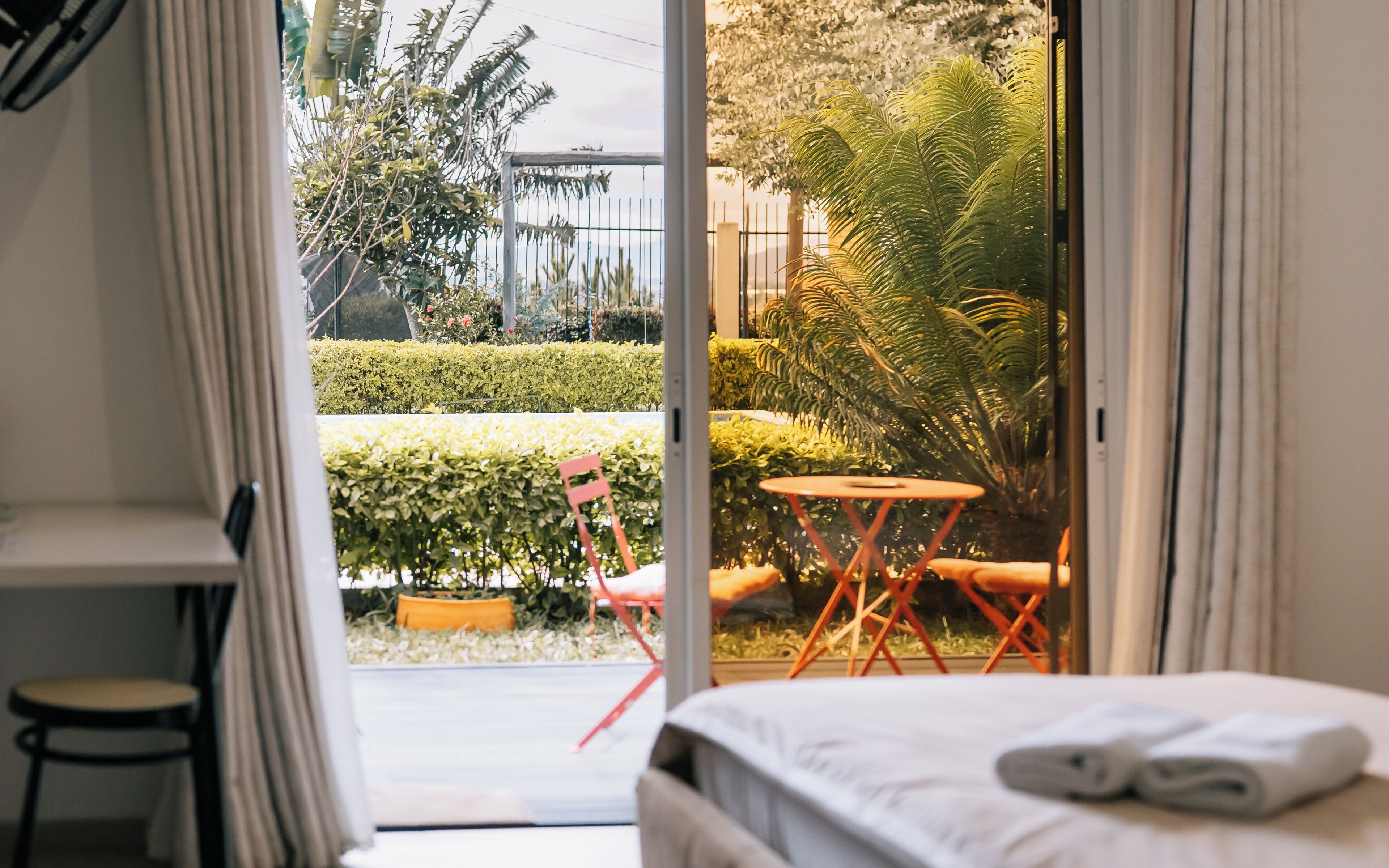 Habitación de hotel con una cama, una pequeña mesa y una silla. Se ve un jardín con seto, una palmera y un edificio al fondo.