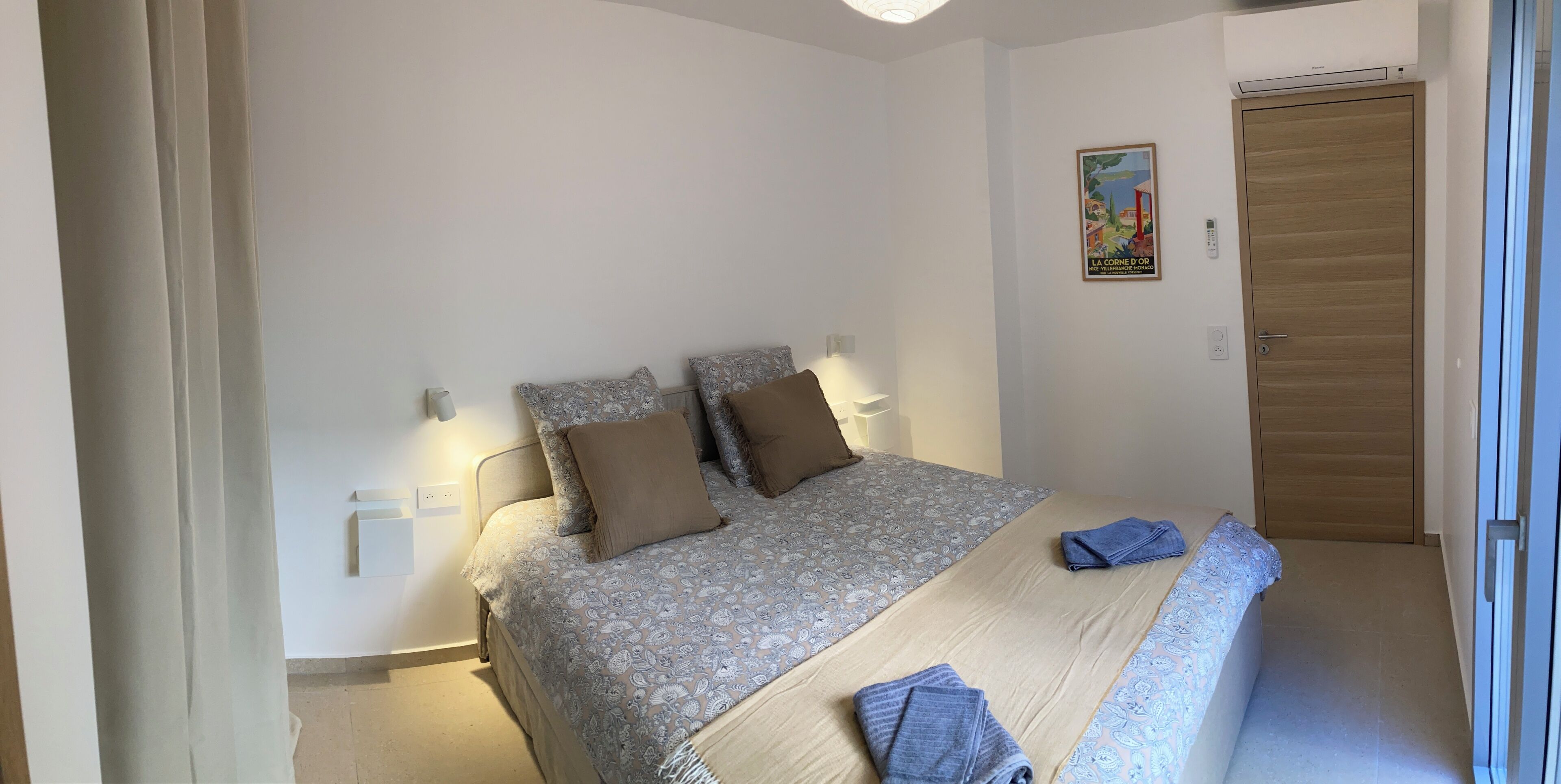 A bedroom with a bed, pillows, a bedside table, a wall-mounted light, a door, and a poster on the wall.