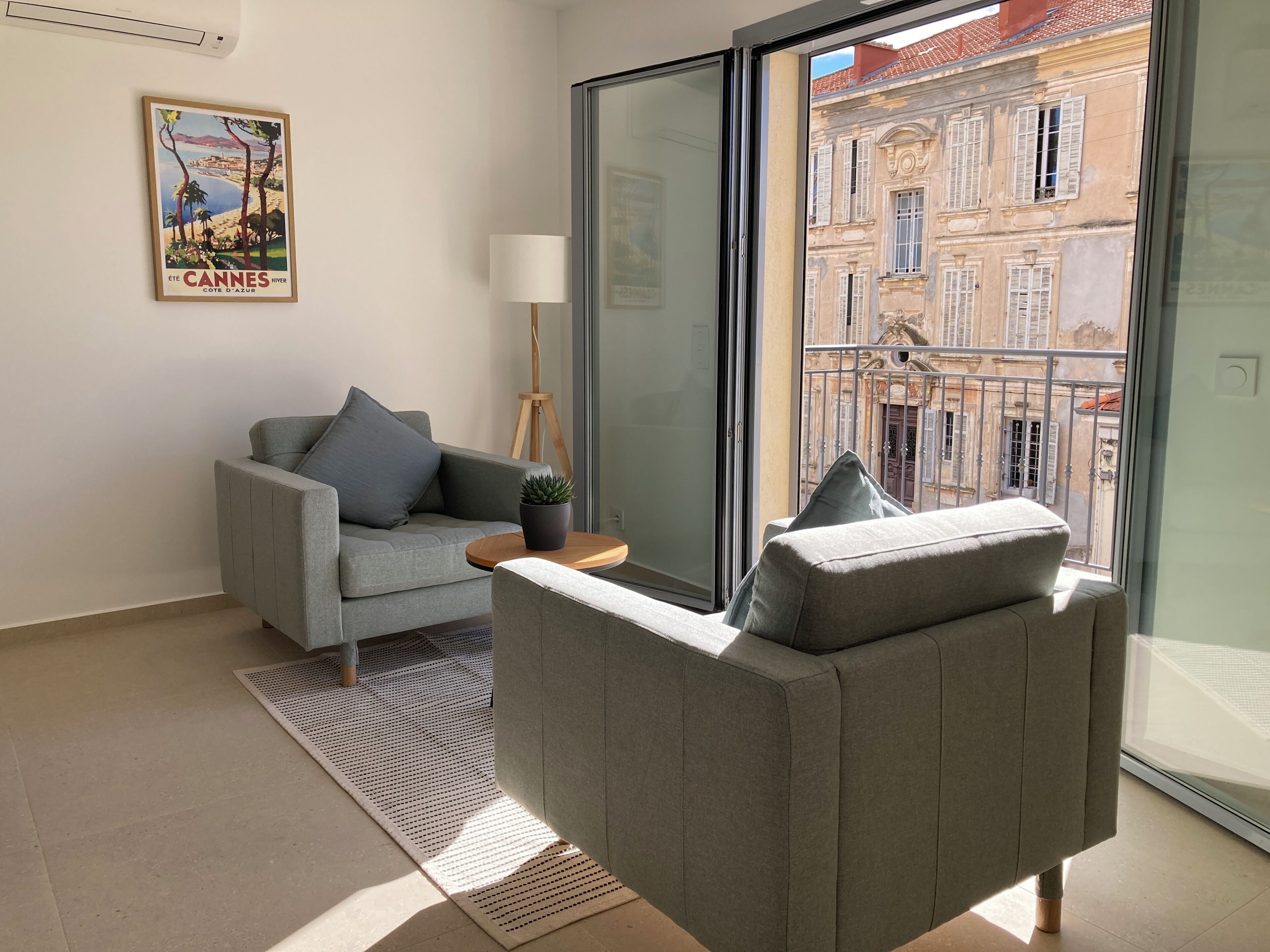 Un soggiorno moderno con un divano grigio, un tavolino e una pianta in vaso. Un'ampia finestra offre vista su un edificio con balconi.