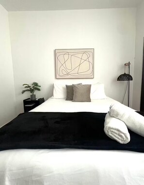 A bedroom with a bed, pillows, a black throw blanket, a white towel, a plant, a floor lamp, and a wall art piece. - Steps to Condado Beach & Ashford Ave (San Juan)
