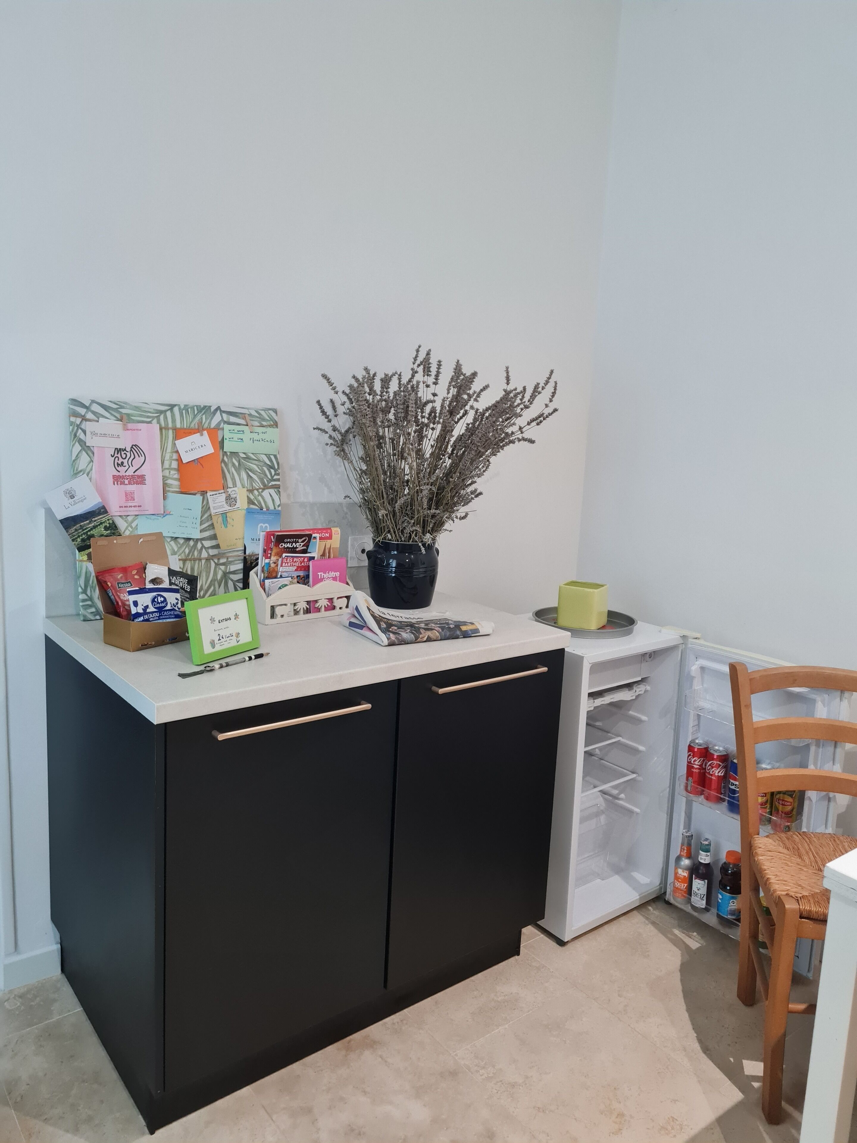 A kitchen area with a white countertop, black cabinets, a potted plant, and various items including a coffee maker, a box of tissues, and a small table with a chair.