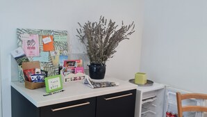 Keittiöalue, jossa on valkoinen työtaso, mustat kaapit, ruukkukasvi sekä erilaisia esineitä, kuten kahvinkeitin, paperipyyhkeiden pakkaus ja pieni pöytä tuolilla.
