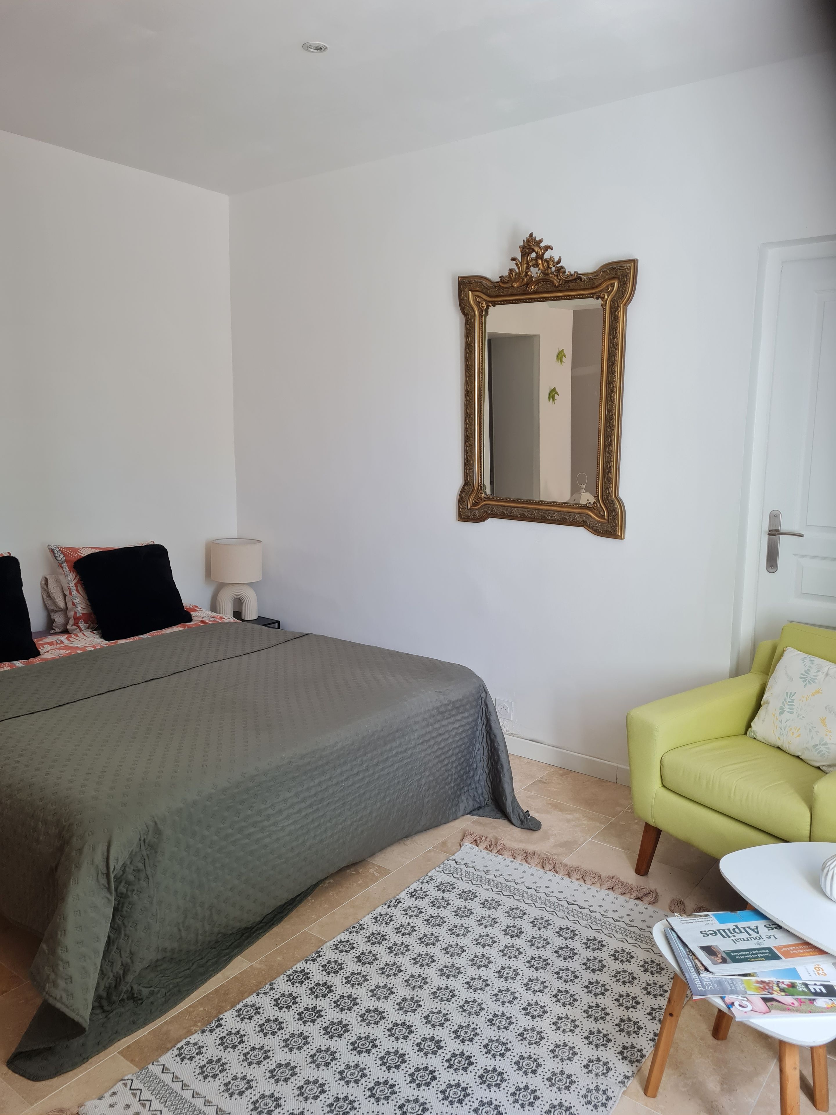 Een slaapkamer met een bed, een stoel, een spiegel en een klein tafeltje met tijdschriften.