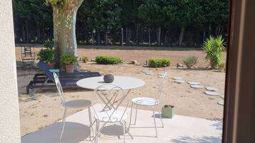 Een terras buiten, met een ronde tafel en stoelen, omgeven door potplanten en een grote boom.
