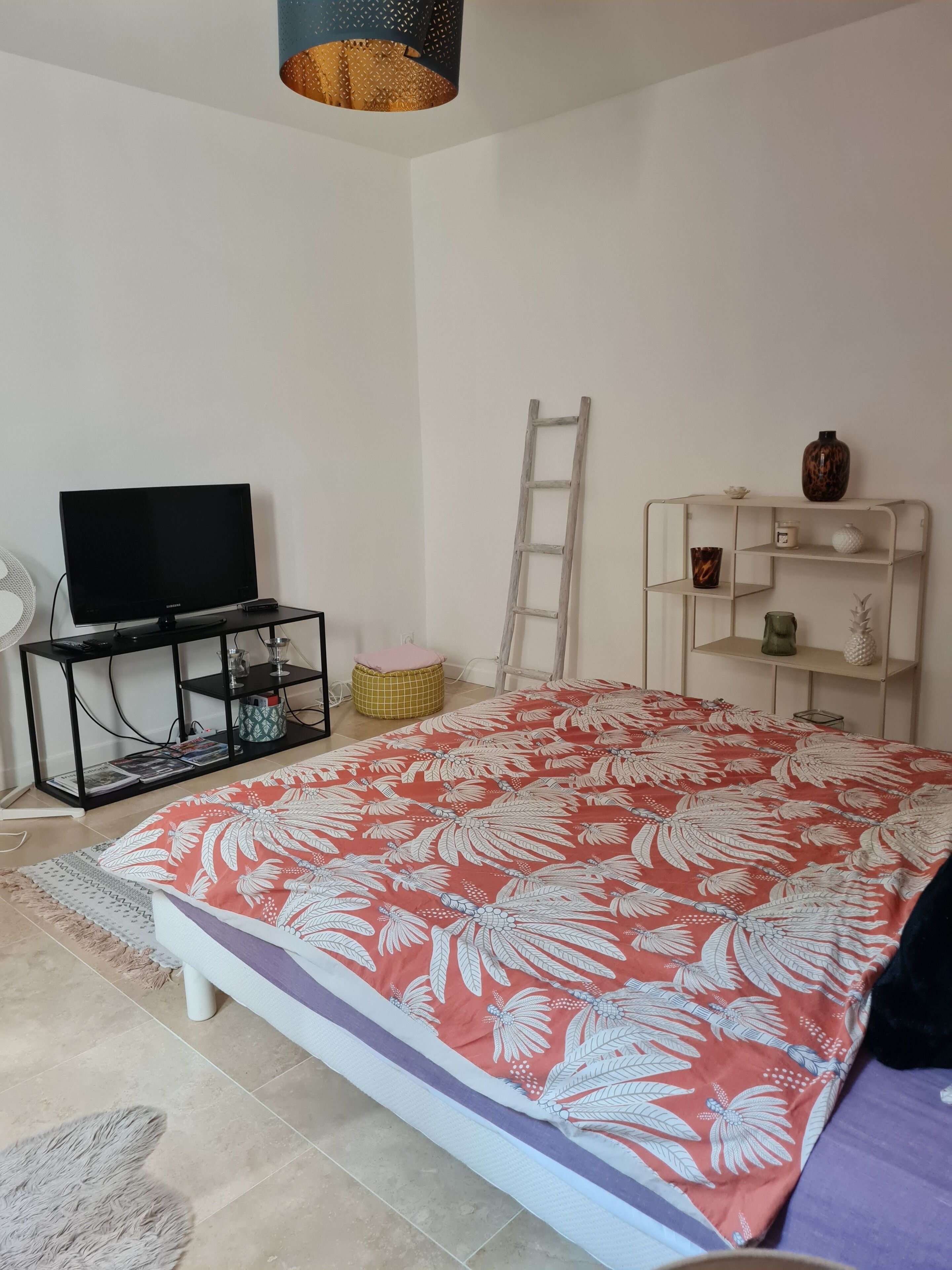 A bedroom with a bed, a TV on a stand, a ladder, a shelf with decorative items, and a fan.