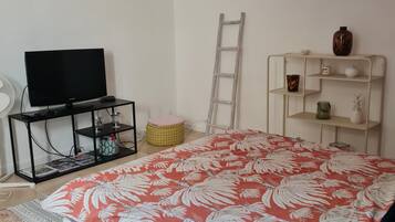 A bedroom with a bed, a TV on a stand, a ladder, a shelf with decorative items, and a fan.
