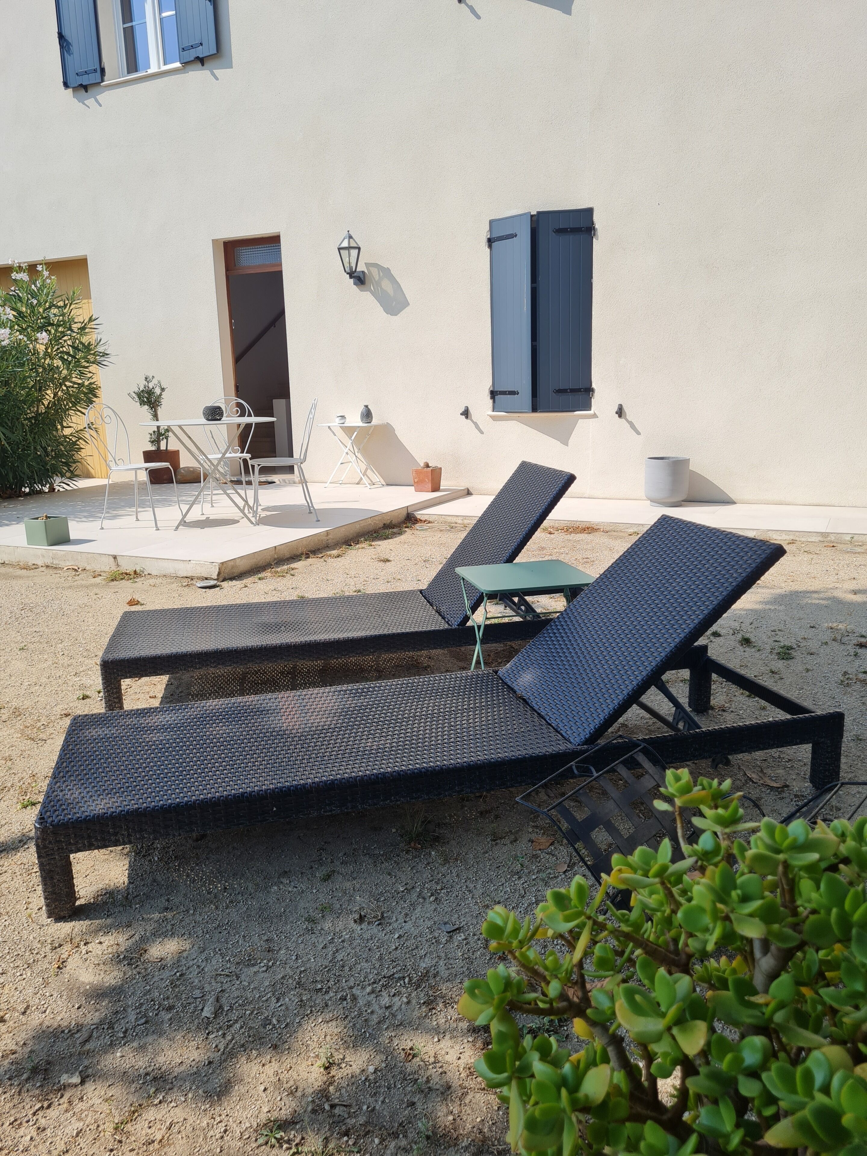 A sunlit outdoor seating area with two lounge chairs and a table, a potted plant, and a building with a door and window.
