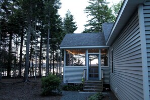 A house with a covered porch, a small front yard, and a forested backdrop. - Romantic Waterfront Cottage with Private Beach & Fireplace Near Hancock, Maine (Hancock)