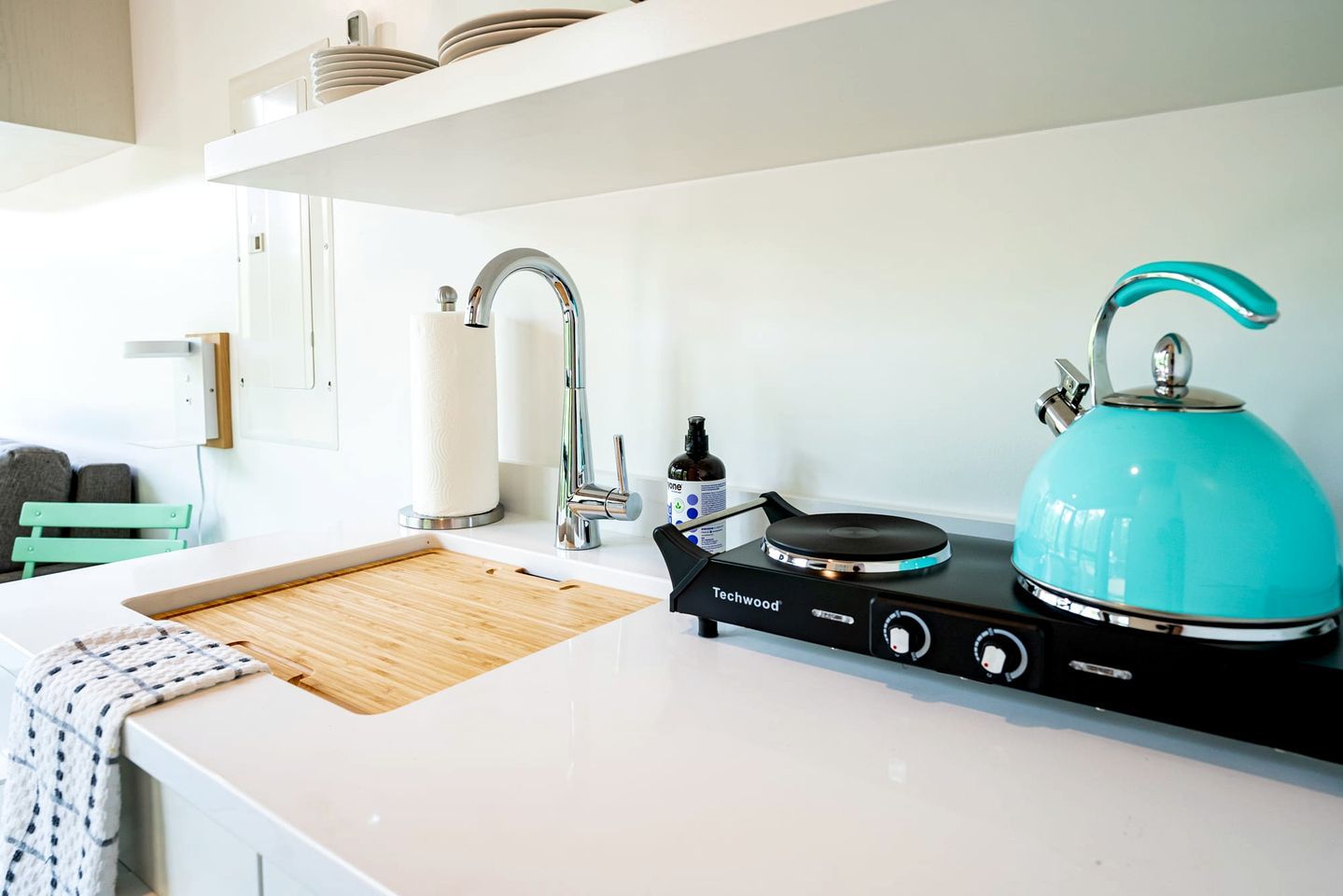 A modern kitchen with a white countertop, a stainless steel faucet, a teal-colored kettle, and a portable gas stove.