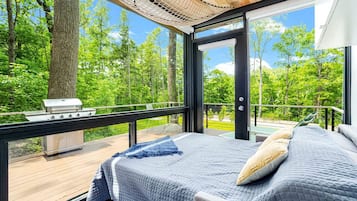Ein Schlafzimmer mit einem großen Bett, Blick auf einen Wald und einem Grill vor der Tür.