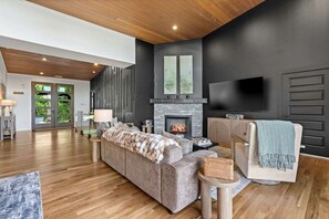 A modern living room with a fireplace, a sofa, a TV, and a large window. - Luxurious Modern Mountain Retreat with Hot Tub & Stargazing Dome Near Berkeley Springs, Hedgesville WV (Hedgesville)