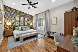 Um quarto com cama, mesas de cabeceira, escrivaninha, cadeira, ventoinha no teto, janela com cortinas e parede decorada com quadros.