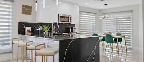A modern kitchen with a central island, bar stools, a dining area with green chairs, and a large window with blinds.